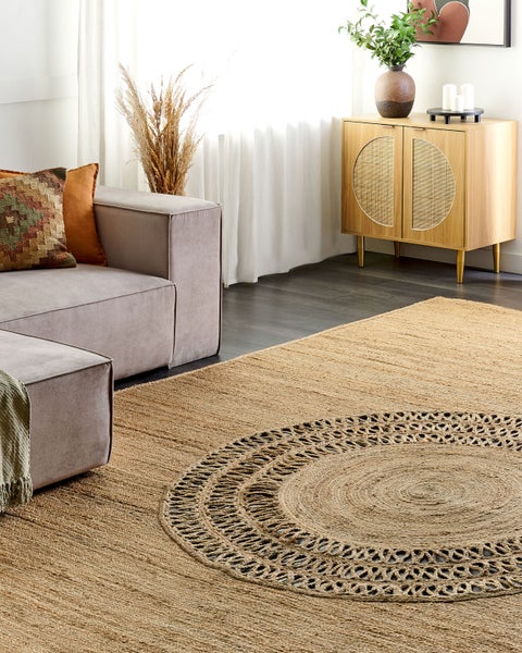 Naturbelassener Jute-Teppich mit zentralem geflochtenem Rundmuster in einem hellen Wohnzimmer mit grauem Sofa und Holz-Sideboard.