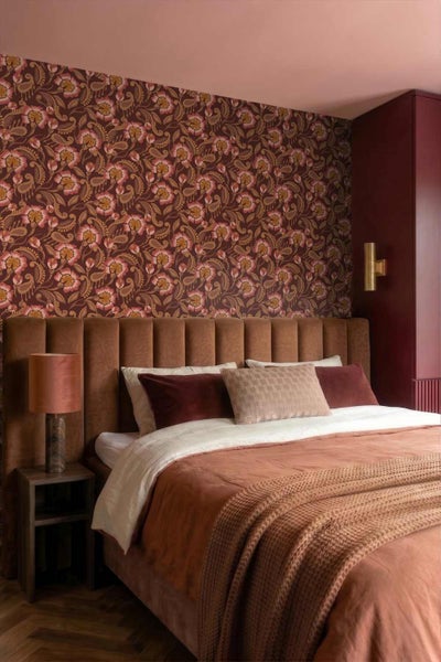 Gemütliches Schlafzimmer mit floral gemusterter Tapete, Bett mit gepolstertem Kopfteil, dekorativen Kissen und einer Lampe auf dem Nachttisch.