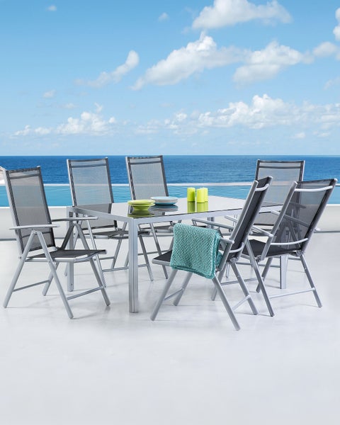 Modernes Gartenmöbel-Set aus grauem Aluminium mit Glastisch und sechs Klappstühlen mit Textilene-Bezug auf einer hellen Terrasse am Meer.