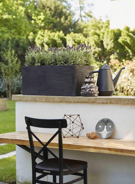 Rechteckiger Pflanzkasten mit strukturierter Oberfläche auf einer Gartenmauer, bepflanzt mit Lavendel, neben einer Gießkanne.