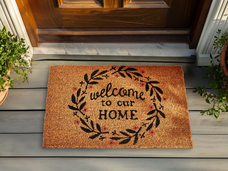Fußmatte aus Kokosfasern mit dem Schriftzug welcome to our home und einem Kranz vor einer Haustür auf einer Holzterrasse.