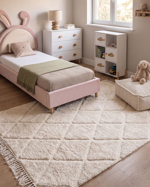 Einrichtung eines Kinderzimmers mit einem Bett mit Hasenohren Kopfteil, einer Kommode, einem Regal und einem hochflorigen Teppich mit Rautenmuster.