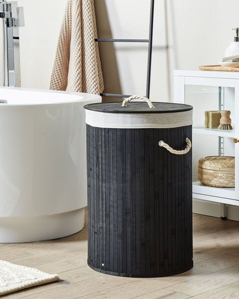 Runder Wäschesammler aus schwarzem Bambus mit Seilgriffen in einem hellen Badezimmer neben einer weißen Badewanne und einer schwarzen Handtuchleiter.