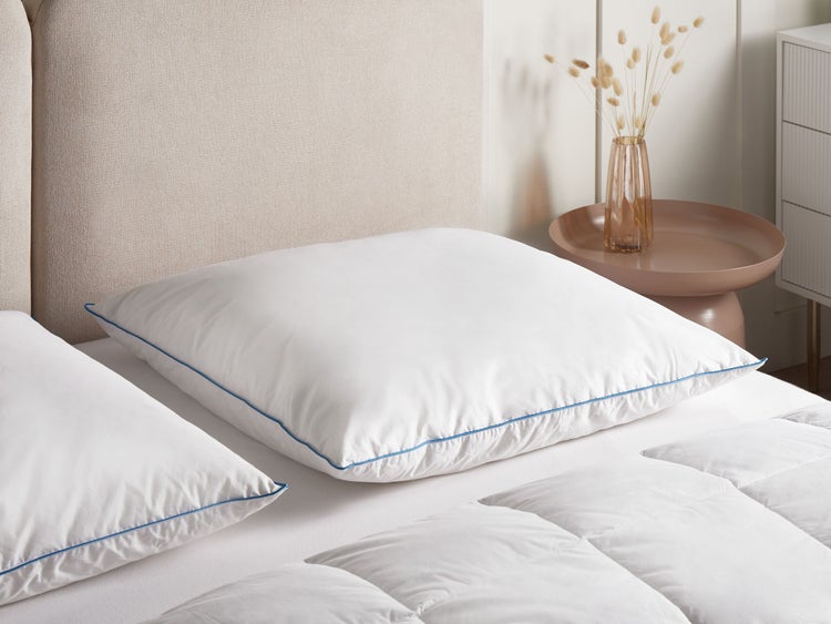 Helles Schlafzimmer mit zwei weißen quadratischen Kopfkissen mit blauer Paspelierung, einer weißen Bettdecke und einem neutralen Kopfteil.