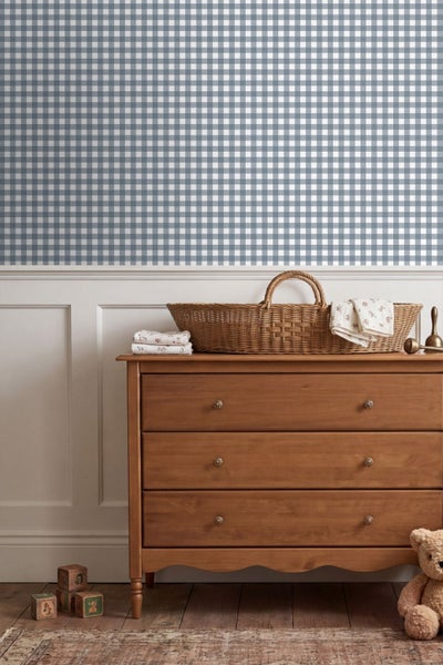 Vliestapete mit blau-weißem Karomuster in einem hellen Kinderzimmer über einer weißen Wandpaneele und einer Holzkommode.