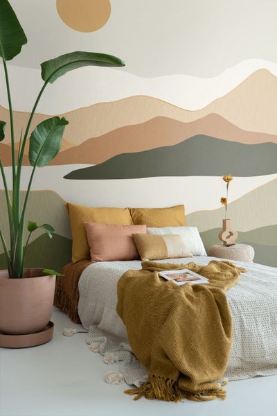 Modernes Schlafzimmer mit Wandbild in Berglandschaft-Optik, Bett mit Kissen und Decke sowie einer großen Zimmerpflanze im Topf.