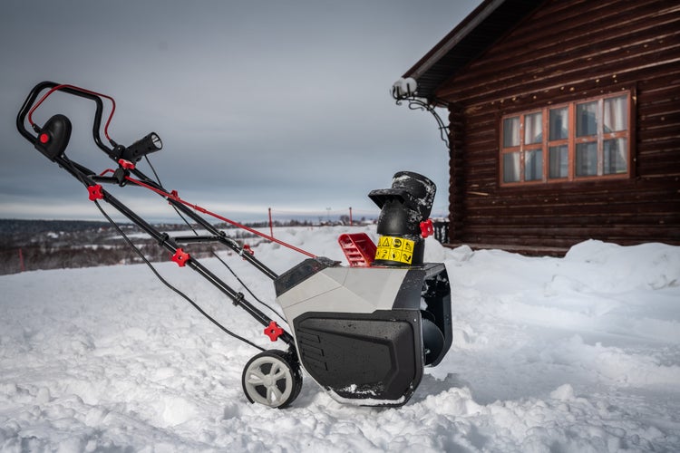 Elektro-Schneefräse auf einer verschneiten Fläche vor einer Holzhütte im Winter.