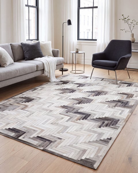 Modernes Wohnzimmer mit einem Teppich aus Fellstücken in Fischgrätenmuster vor einem grauen Sofa und einem Sessel auf Parkettboden.