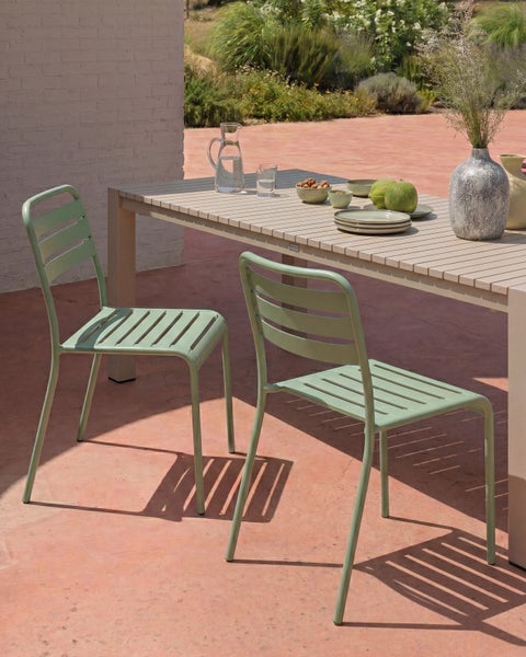 Garten-Essgruppe mit einem rechteckigem Lamellentisch und zwei grünen Metallstühlen auf einer sonnigen Terrasse vor einer weißen Backsteinmauer.