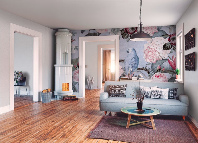 Helles Wohnzimmer mit großflächiger Blumentapete mit Papagei-Motiv, Sofa, Holzcouchtisch und Kachelofen auf Echtholzboden.