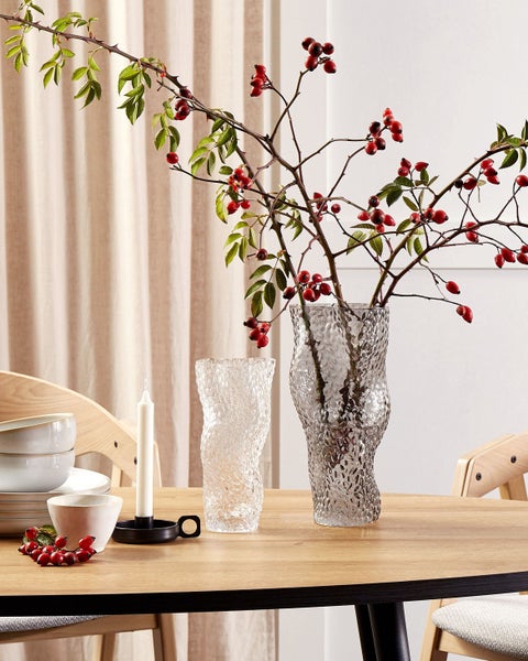 Tischdekoration mit zwei Glasvasen und Hagebuttenzweigen auf einem Holztisch.