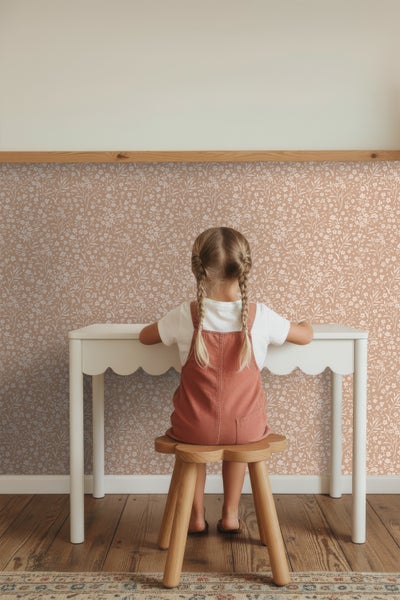 Mädchen sitzt an weißem Schreibtisch vor gemusterter Tapete auf einem Hocker