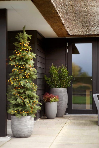 Terrasse mit strukturierten Pflanzkübeln und einem beleuchteten Weihnachtsbaum in einem Topf vor einer dunklen Holzfassade.