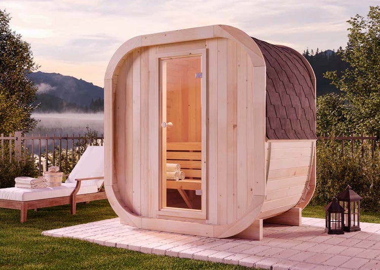Moderne Fasssauna aus Holz für den Garten mit Glastür und Schindeldach auf gepflastertem Untergrund in landschaftlicher Umgebung.