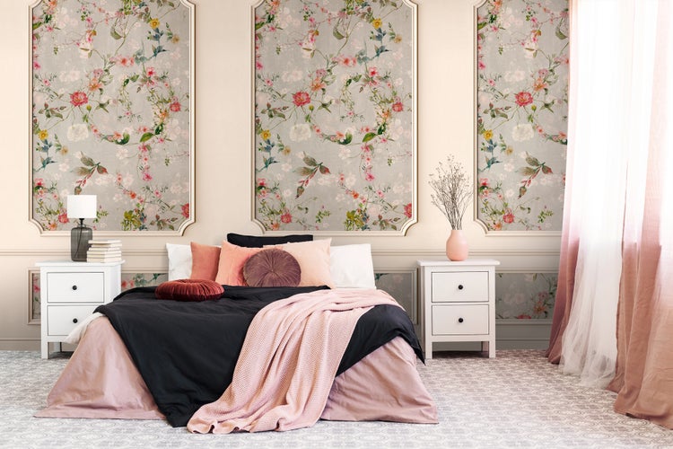 Hell gestaltetes Schlafzimmer mit floralen Tapetenpaneelen in Wandrahmen, einem Bett mit rosa und schwarzer Bettwäsche sowie weißen Nachttischen.