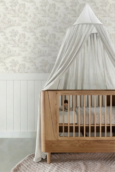 Kinderbett aus hellem Holz mit Betthimmel aus Stoff vor gemusterter Tapete und weißer Wandvertäfelung.