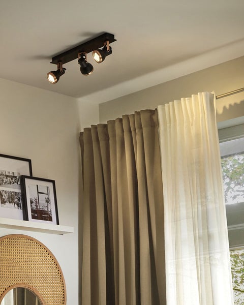 Dreiflammiger schwarzer Deckenspot in einem hellen Raum mit Vorhängen, Wandregal mit Bildern und einem Rattanspiegel.