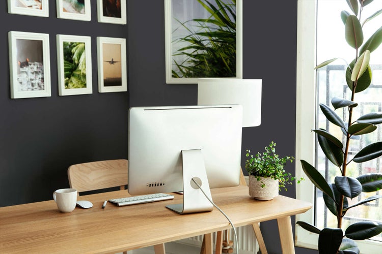 Modernes Arbeitszimmer mit anthrazitfarbener Wand, hellem Holzschreibtisch, Computer und weißen Bilderrahmen.