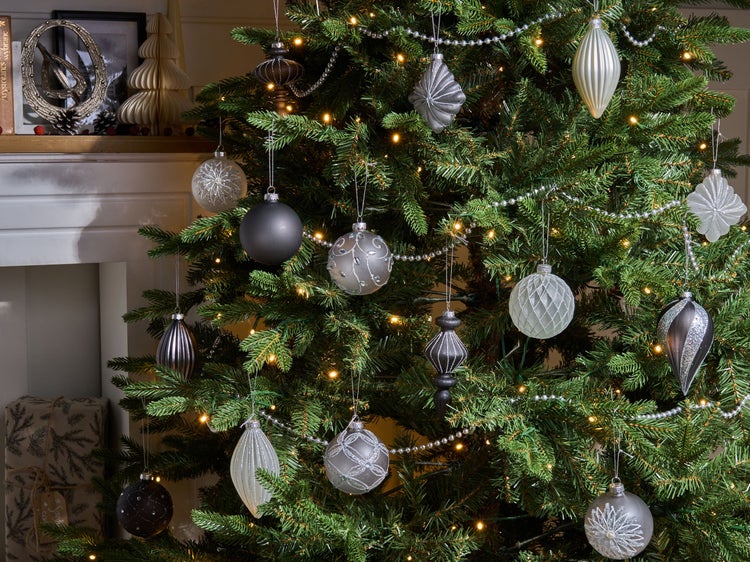 Weihnachtsbaum mit grauen, silbernen und weißen Kugeln, silberner Perlenkette und warmweißer Lichterkette vor einem Kamin.