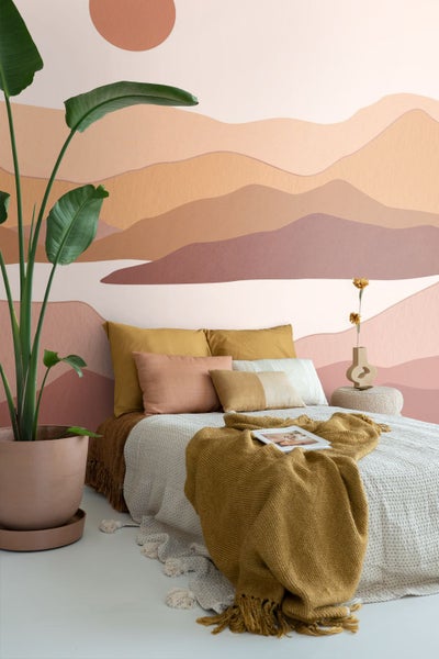 Fototapete mit abstrakter Berglandschaft in Erdtönen in einem modern eingerichteten Schlafzimmer mit Bett und einer großen Zimmerpflanze.
