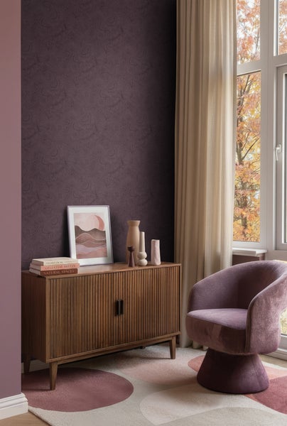 Wohnzimmer mit dunkelvioletter Mustertapete, Sideboard aus Holz, einem lila Sessel und hellen Vorhängen neben einem Fenster.
