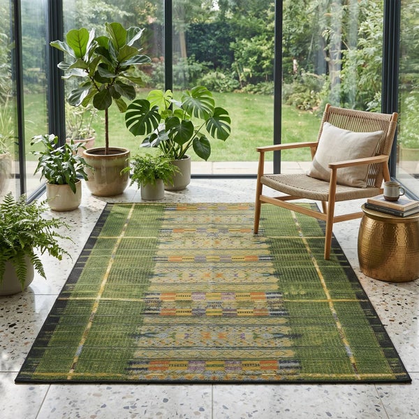 Hell eingerichteter Raum mit grün gemustertem Teppich, Holzstuhl, Goldtisch und Zimmerpflanzen vor einer Glasfront mit Blick in den Garten.