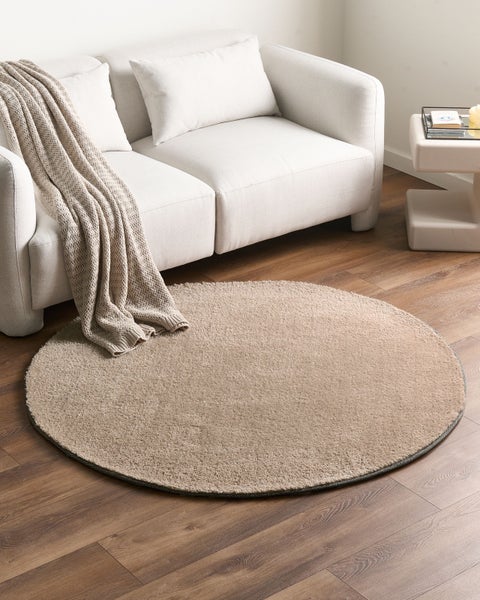 Runder, flauschiger beigefarbener Teppich auf einem dunklen Holzboden vor einem hellen Sofa in einem hellen Wohnzimmer.