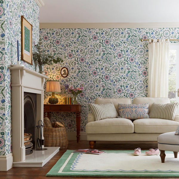 Klassisch eingerichtetes Wohnzimmer mit floral gemusterter Tapete an den Wänden, einem Kamin, einem hellen Sofa und einem Teppich.