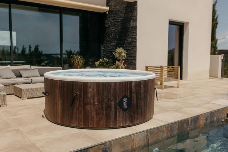 Runder Whirlpool in Holzoptik auf einer hellen Terrasse neben einem Pool, mit Loungemöbeln und Coco Spa Logo.