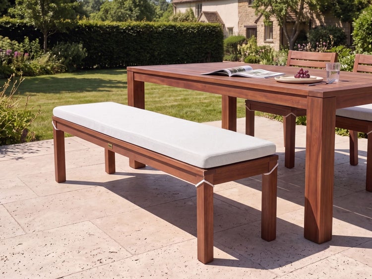 Gartenbank aus Holz mit Auflage auf einer Terrasse mit Gartentisch und Stühlen vor einem Haus.
