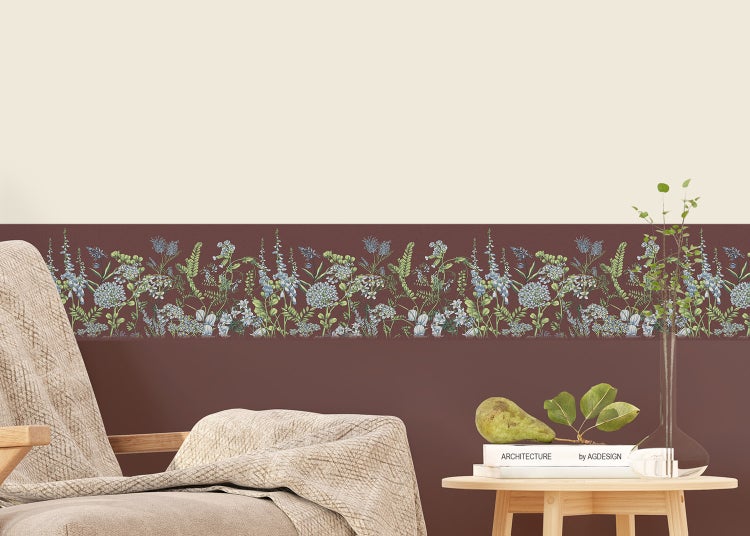Tapetenbordüre mit Wiesenblumen-Motiv an einer zweifarbigen Wand in einem hellen Wohnraum mit Sessel und Holztisch.