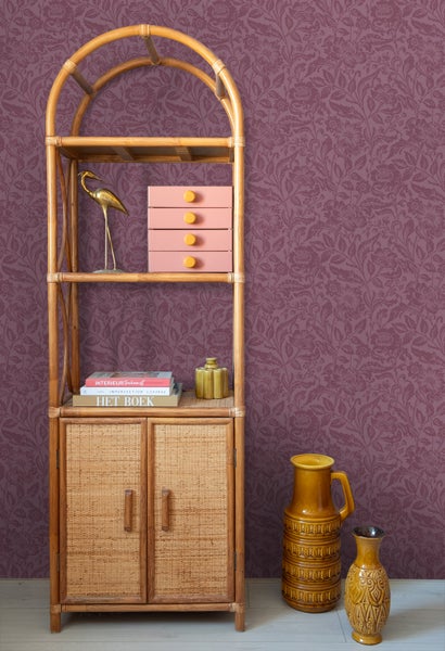 Szene mit einem Regalschrank aus Rattan, dekoriert mit Accessoires vor einer floralen Tapete.
