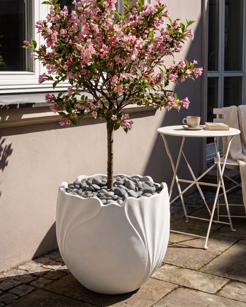Dekorativer weißer Pflanztopf in Kelchform mit blühendem Stämmchen und Kieselsteinen auf einer Terrasse mit Bistrotisch.