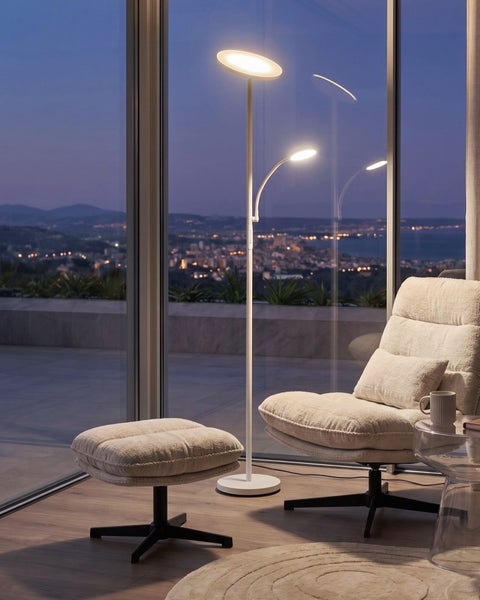 Moderne Stehlampe mit Deckenfluter und Leselicht neben einem bequemen Sessel mit Fußhocker vor einer großen Fensterfront mit Stadtblick bei Nacht.