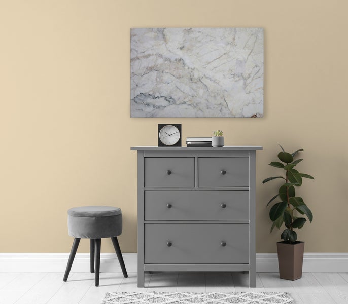 Eine Kommode mit vier Schubladen steht vor einer Wand mit einem Marmor Wandbild, ergänzt durch einen Hocker, eine Pflanze und eine Tischuhr.