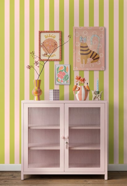 Hell gestalteter Raum mit rosa Schrank mit geriffelten Glastüren vor gestreifter Tapete, dekoriert mit Vasen und Wandbildern.
