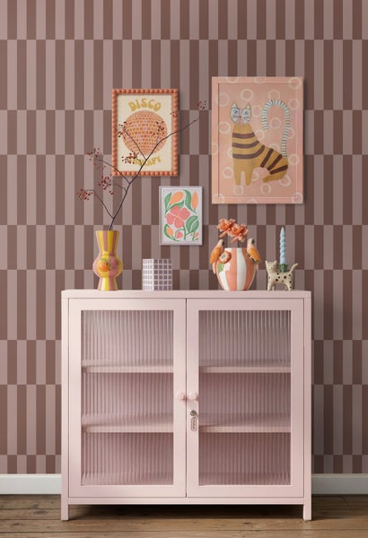Rosa Metallschrank mit geriffelten Glastüren vor geometrisch gemusterter Tapete mit bunten Vasen und Wandbildern.