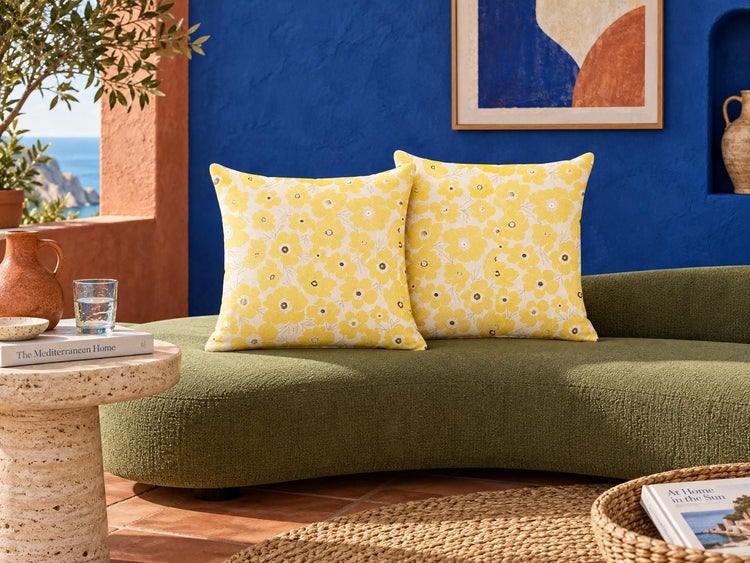 Zwei quadratische Kissen mit Blumenmuster auf einem geschwungenen Sofa in einem mediterran eingerichteten Raum.