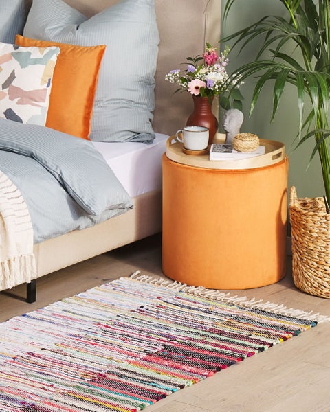 Helles Schlafzimmer mit einem orangefarbenen Samthocker als Beistelltisch, einem bunten gewebten Teppich und blauer Bettwäsche.