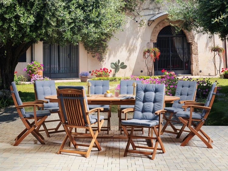 Gartenmöbel-Set mit ovalem Holztisch und sechs Holz-Klappstühlen mit blauen Polstern auf einer gepflasterten Terrasse im sonnigen Garten.