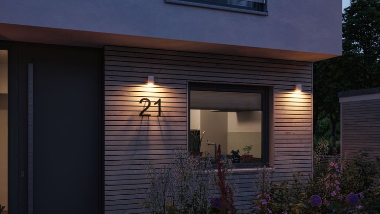Moderne Außenwandleuchten beleuchten eine Holzfassade mit der Hausnummer 21 neben einem Fenster bei Dämmerung.