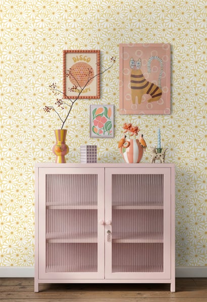 Vliestapete mit weißem Gänseblümchen-Muster auf gelbem Grund, kombiniert mit einer rosa Vitrine in einem hellen, farbenfrohen Raum.