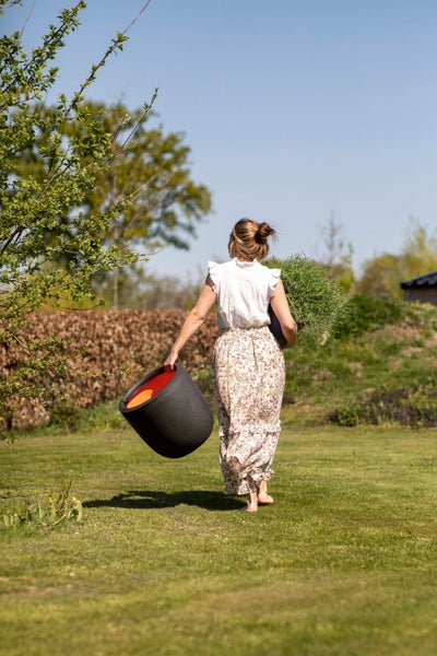 Eine Frau trägt einen großen schwarzen Pflanzkübel und eine Pflanze über eine sonnige Rasenfläche im Garten.