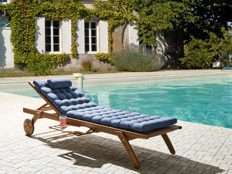 Gartenliege aus Holz mit blauer Auflage und Rollen auf einer hellen Steinterrasse an einem Pool in einem hellen, organisierten Außenbereich.
