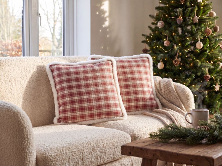 Gemütliches Wohnzimmer mit zwei rot-weiß karierten Kissen mit Kunstfellrand auf einem beigefarbenen Sofa neben einem geschmückten Weihnachtsbaum.