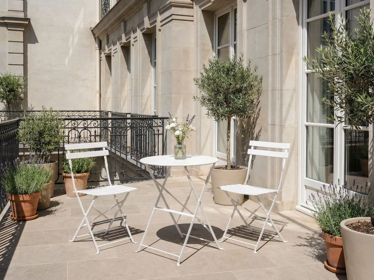 Weiße Bistro-Garnitur aus Metall mit rundem Klapptisch und zwei Klappstühlen auf einer sonnigen Terrasse mit vielen Topfpflanzen.