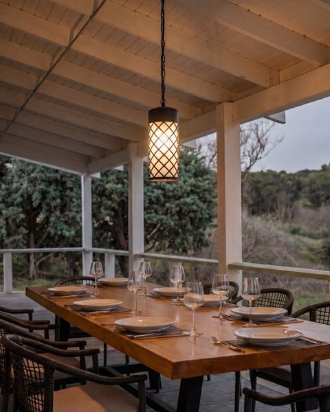 Überdachte Terrasse mit Holzesstisch und Hängeleuchte im Gitterdesign, dazu Stühle aus Korbgeflecht und eine gedeckte Tafel.