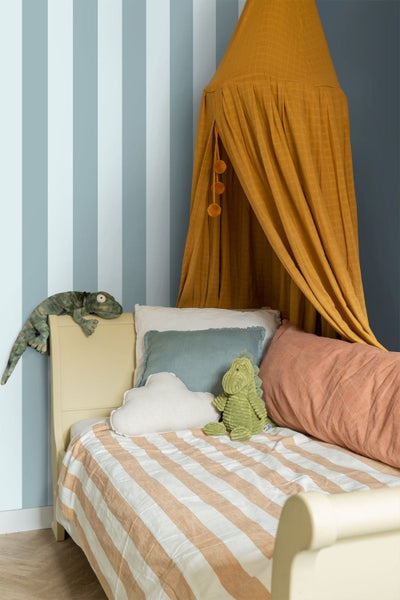 Helles Kinderzimmer mit blau-weiß gestreifter Vliestapete, einem Holzbett mit Stoff-Betthimmel, verschiedenen Kissen und weichen Kuscheltieren.