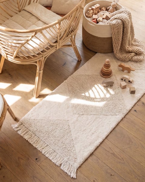 Ein Kinderzimmer mit einem Rattan Sessel, einem strukturierten Teppich mit Fransen und einem Korb voller Holzspielzeug auf einem Holzboden.