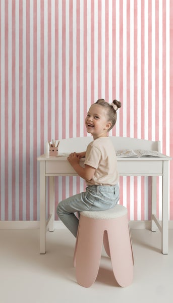 Mädchen sitzt an Schreibtisch auf Hocker vor gestreifter Tapete